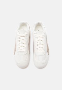 Par belih Puma suede športnih copat z svetlo rjavimi stranskimi črtami, zvezanih, videnih od zgoraj na enobarvni beli podlagi.