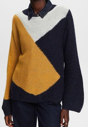Osoba nosząca gruby sweter z dużymi geometrycznymi blokami w kolorach musztardowej żółci, granatu i bieli, w połączeniu z ciemnymi dżinsami.