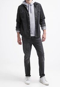 Mężczyzna w szarej bluzie z kapturem pod czarną koszulą zapinaną na guziki, w ciemnoszarych dżinsach i białych trampkach stojący na białym tle.