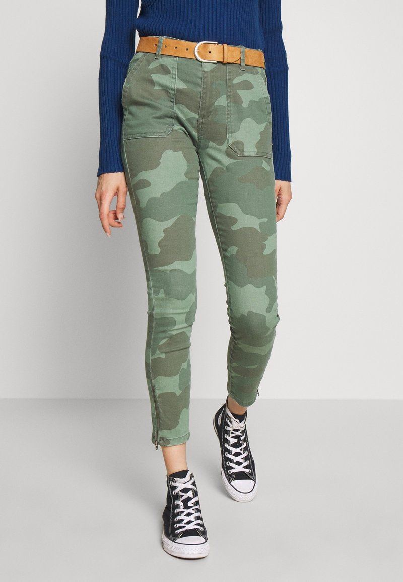 Persona vestida con pantalones de camuflaje verdes con bolsillos frontales, cinturón marrón, suéter azul marino y zapatillas negras de caña alta caminando hacia adelante.
