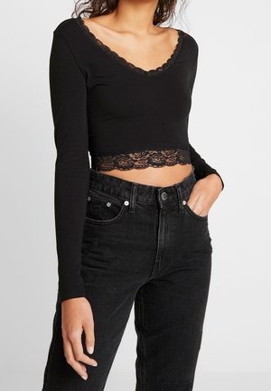 Frau trägt ein schwarzes, langärmeliges Crop-Top mit Spitzenbesatz am Ausschnitt und Saum, kombiniert mit hoch taillierten schwarzen Jeans, Hand in der Tasche.