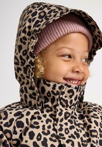 Giacca beige con stampa leopardata, dotata di un alto colletto e di un cappuccio, con dettagli neri. Il bambino indossa un berretto di maglia rosa chiaro sotto.