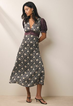 Kvinde med langt mørkt hår iført en marineblå blomster midi kjole med pufærmer og brune sandaler med hæl, stående mod en lys væg.