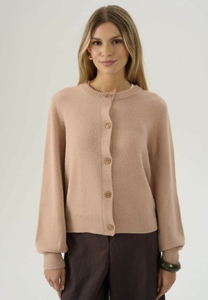 Woman wearing a beige buttoned cardigan with long sleeves and dark brown pants, standing against a plain light background.