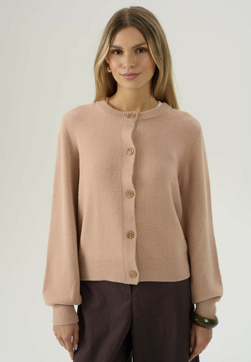 Donna che indossa un cardigan beige con bottoni a maniche lunghe e pantaloni marrone scuro, in piedi davanti a uno sfondo chiaro semplice.