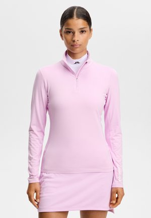 Woman wearing a fitted light pink long-sleeve top with a quarter zip and matching skirt, standing against a plain light background.