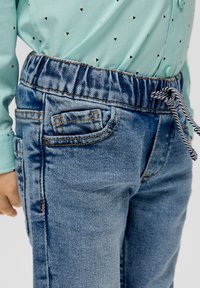 Modri denim kavbojke z elastičnim pasom, ki vključujejo majhen sprednji žep in subtilne detajle bledenja. V kompletu z zeleno-mint vzorčeno majico.