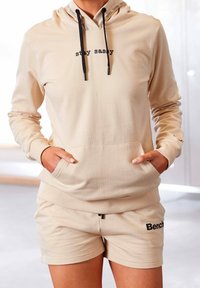 Sudadera con capucha beige con bolsillo canguro y cordones, con el texto "stay sassy" en el frente. Combinada con pantalones cortos beige a juego.