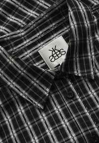 Melns un pelēks piparu raksts ar pogām uz augšu, izplatītā apkakle un balts zīmola marķējums apakšā apkakles iekšpusē.