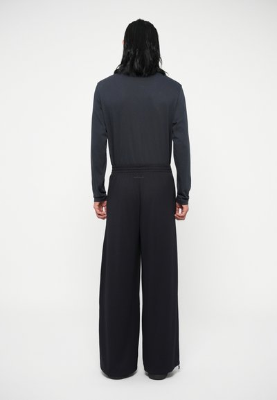 Ilgesniam rankovėms tamsiai pilkas marškinėliai derinami su plačiomis juodomis kelnėmis, turinčiomis tekstūruotą medžiagą ir elastinį juosmenį, matomi iš nugaros.