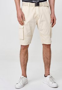 Beige Cargo-Shorts mit zwei seitlichen Taschen, genähten Details und einem umgeschlagenen Saum. Kombiniert mit weißen Sneakers und einem schwarzen Taillengummi.