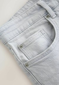 Poche de jean en denim gris clair avec passants de ceinture, rivets métalliques et détails de couture visibles sur la texture du tissu.
