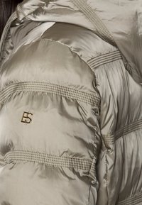 Giacca imbottita in taupe metallizzato con texture trapuntata, caratterizzata da cuciture orizzontali e un logo dorato sulla manica. Finitura liscia e lucida.