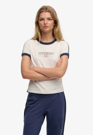Cremefarvet t-shirt med marineblå kant og korte ærmer, med "SUPERDRY" grafik og logo på forsiden. Kombineret med marineblå joggingbukser.