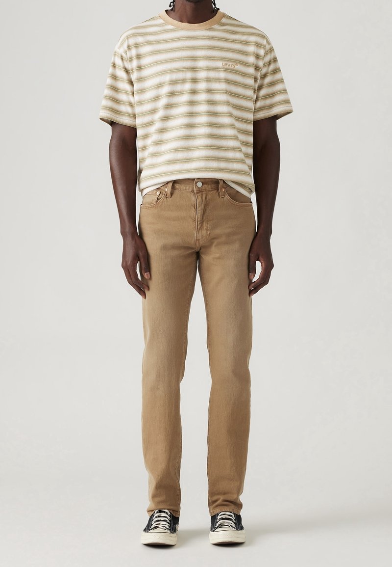 Camiseta a rayas en beige y blanco con cuello redondo y logo, combinada con jeans ajustados de color marrón claro y zapatillas negras con suelas blancas.