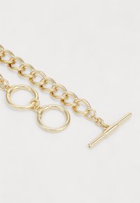 Gullfarget lenke armbånd med ovale ledd, to sirkulære ringer og en T-bar lås for sikker festing. Glatt metallisk overflate.
