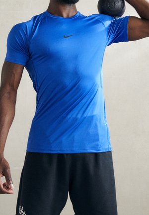 Homme portant un maillot de sport bleu Nike et un short noir, tenant une kettlebell reposant sur son épaule devant un fond uni.