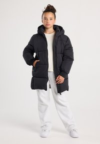 Chica que lleva una chaqueta acolchada con capucha negra, una camiseta blanca, pantalones de chándal grises y zapatillas blancas, de pie con una mano en el bolsillo.