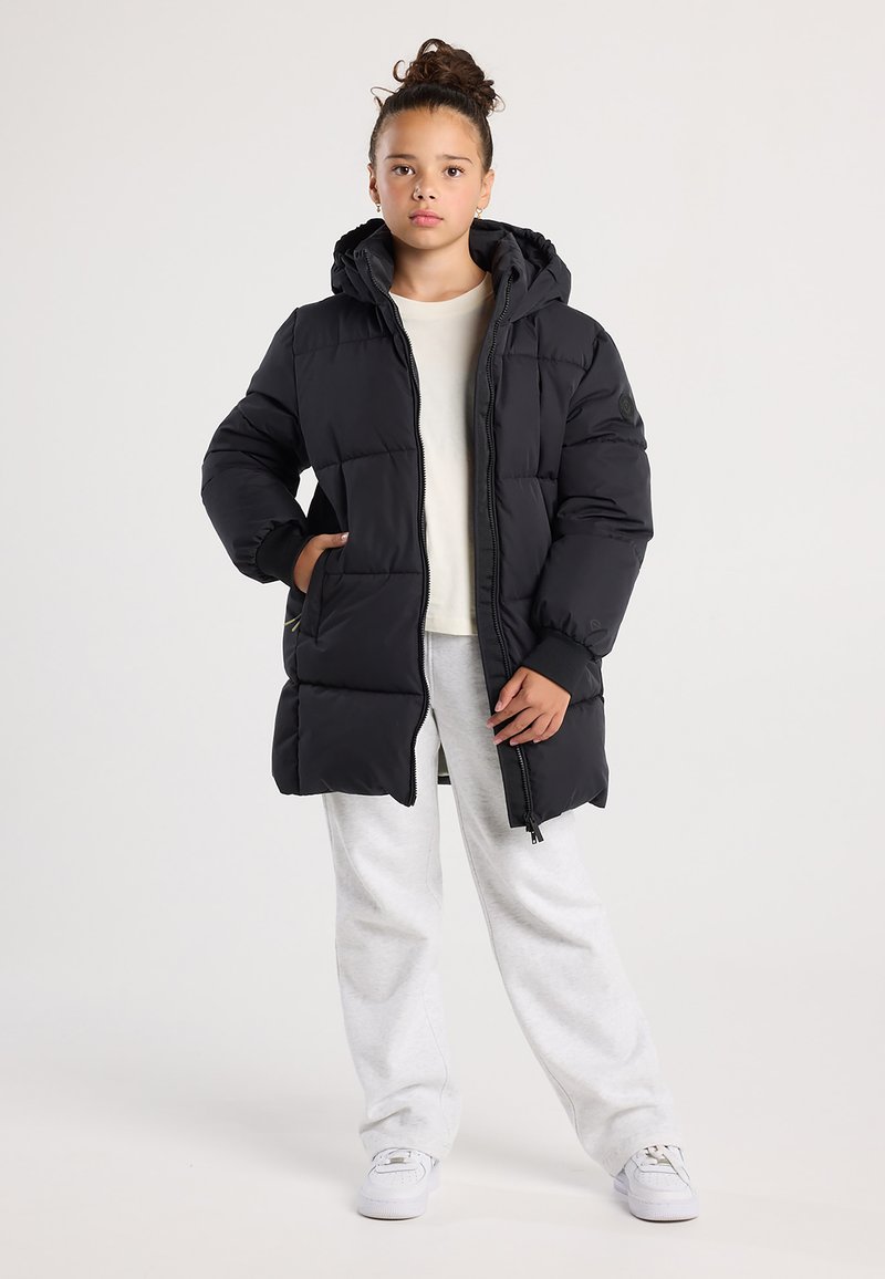 Chica que lleva una chaqueta acolchada con capucha negra, una camiseta blanca, pantalones de chándal grises y zapatillas blancas, de pie con una mano en el bolsillo.