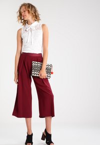 White lace blouse with a collar and bow detail, burgundy wide-leg trousers, black open-toe heeled sandals, and a patterned clutch bag.