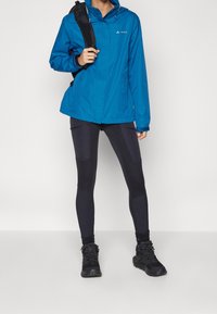 Veste imperméable bleue avec capuche, poignets réglables et deux poches avant. Associée à des leggings noirs avec des panneaux texturés et des chaussures noires.