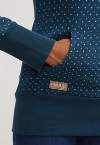 Main dans la poche avant d'un sweat-shirt bleu à pois avec une étiquette de la marque Ragwear sur la poche.