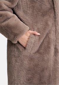 Hånd i lommen på en blød, luksuriøs lysebrune faux fur frakke vist fra taljen og ned.
