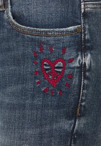 Vaqueros de mezclilla con un lavado azul oscuro desgastado que presenta un corazón bordado en rojo y un diseño de sol radiante en el bolsillo trasero. Estilo clásico de cinco bolsillos.