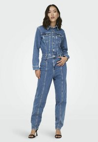 Une femme se tient debout, portant une veste en jean bleue et un jean taille haute assorti avec des coutures verticales, accompagnés de talons noirs ouverts à l'avant.