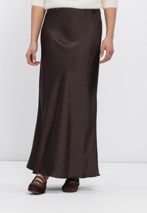 Femme portant une longue jupe en satin marron foncé avec des ballerines marron à lacets et un pull crème, debout devant un fond blanc uni.