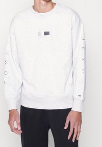 Sudadera de algodón gris con un parche de logo rectangular en blanco y negro, con "TOMMY JEANS" impreso a lo largo de las mangas. Puños acanalados.