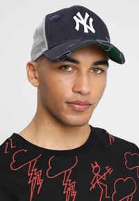 Marineblaue Baseballmütze mit weißem Logo, ausgefransten Kanten und grauem Netzrücken. Die Mütze hat ein grünes Untervisier und eine weiche Textur.