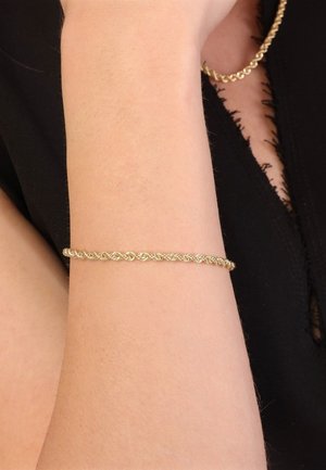 Goldfarbener Armband mit einem gedrehten Design, das eine glatte Oberfläche und eine runde Form aufweist, getragen an einem nackten Handgelenk vor einem dunklen Hintergrund.