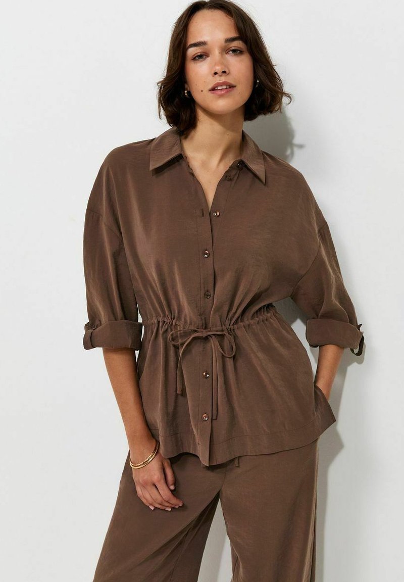 Woman with short dark hair wearing a brown button-up shirt with rolled sleeves and matching brown pants against a neutral background.