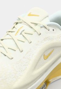 Nike-schoenen met een lichtcrème bovenkant, textuur overlays, gele accenten en een transparante zool voor verbeterde zichtbaarheid.
