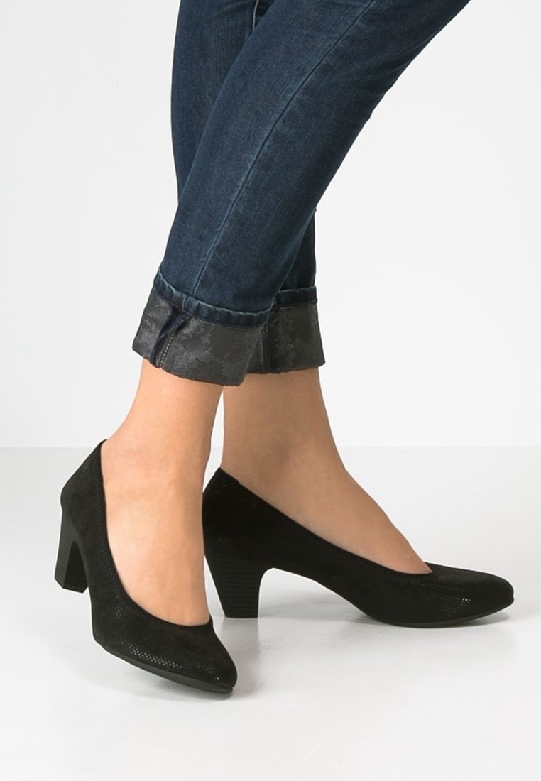 Femme portant un jean skinny bleu foncé retroussé et des chaussures à talons bas noirs sur un fond clair uni.
