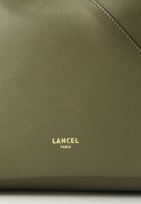 Olivinės spalvos odinė rankinė su lygiu paviršiumi, pažymėta auksinėmis raidėmis "LANCEL PARIS" apačioje, su įstrižais siuvinėjimo detalėmis.