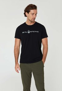 Černé bavlněné tričko s krátkým rukávem, na přední straně se nachází bílé logo "SAIL RACING". Model je spojený s olivově zelenými kalhotami.