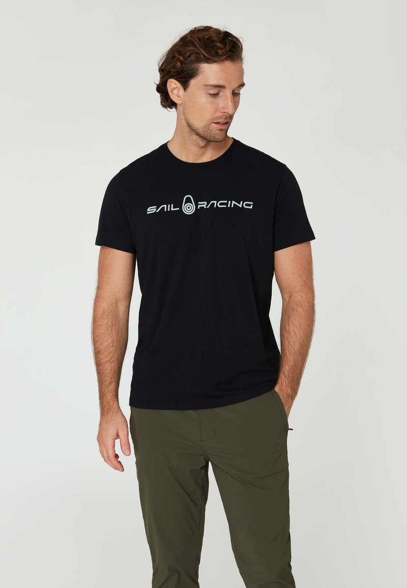 Černé bavlněné tričko s krátkým rukávem, na přední straně se nachází bílé logo "SAIL RACING". Model je spojený s olivově zelenými kalhotami.