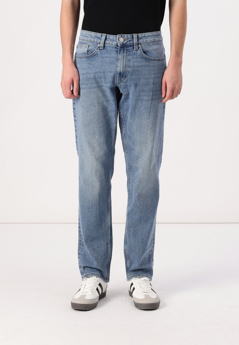 Lyseblå denimjeans med rett snitt, fem lommer og synlige sømdetaljer, brukt med hvite sneakers med svarte akcenter.