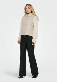 Hellbeige Steppweste mit hohem Kragen, horizontaler Steppung und Frontreißverschluss, kombiniert mit schwarzen ausgestellten Hosen und spitzen schwarzen High Heels.