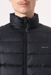 Juoda pūkinė striukė su aukšta apykakle, vertikaliu siūlumų raštu, priekiniais užtrauktuku ir subtiliu logotipu kairiajame krūtinės regione.