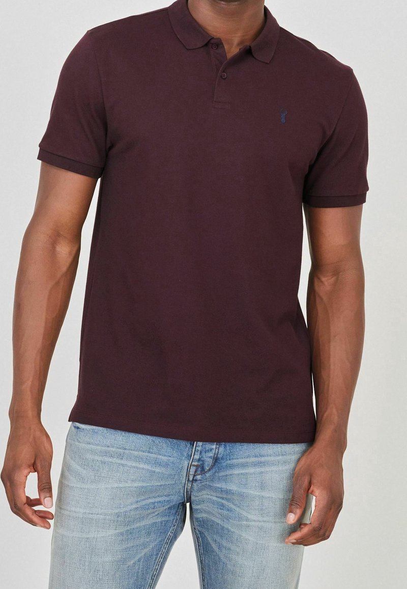 Homme portant un polo manches courtes de couleur bordeaux foncé et un jean bleu clair, debout avec les bras détendus le long du corps.
