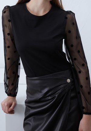 Top noir avec manches bouffantes en tulle à pois, associé à une jupe en similicuir noir avec un pli diagonal et un bouton métallique.