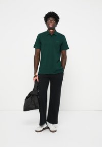 Camisa polo verde de manga curta com um logótipo discreto, combinada com calças largas pretas e ténis brancos, segurando uma mala acolchoada preta.