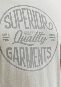 Hellgraues Baumwoll-T-Shirt mit einem großen runden Grafikelement, auf dem der Text "SUPERIOR QUALITY GARMENTS" in Dunkelgrau steht.