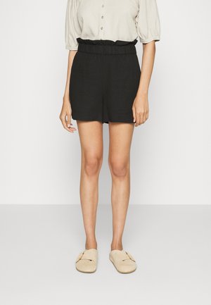 Frau trägt hoch taillierte schwarze Shorts mit gerafftem Bund, beige Bluse mit Knöpfen und beige Slip-On-Schuhe, steht vor weißem Hintergrund.