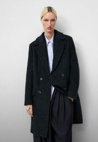 Cappotto in lana verde scuro doppiopetto con ampi revers, dettagli in bottone e finitura texturizzata, abbinato a una camicia azzurra chiara e pantaloni scuri.