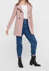 Cappotto in misto lana di color rosa chiaro con un ampio colletto e chiusura doppiopetto, abbinato a jeans blu a vita alta e stivali neri alla caviglia.