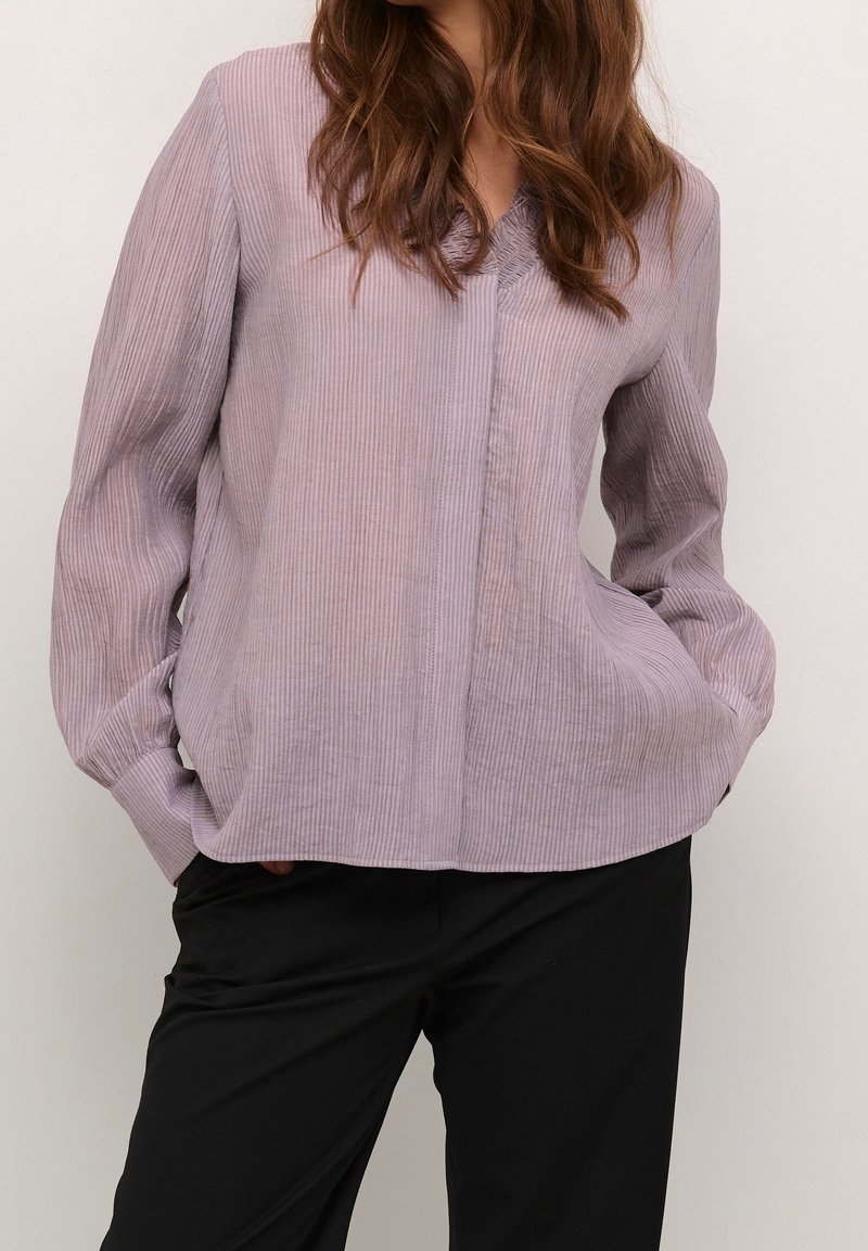 Blouse à manches longues de couleur violet clair avec une texture en côtes verticales. Encolure en V avec de légers plis, poches latérales et coupe ample.
